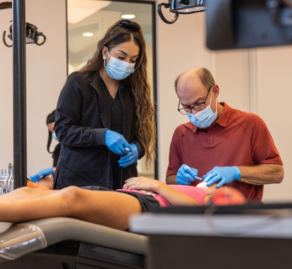 Two healthcare professionals wearing masks and gloves examine a patient's arm in a clinical setting.